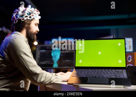 Focus on green screen laptop used by programmer using EEG headset to upload mind into PC. Close up of mockup notebook on desk next to man experimenting with transferring consciousness into cyberspace Stock Photo