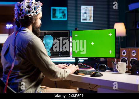 IT technician using EEG headset device translating thoughts into commands on green screen PC display on desk. System administrator controlling chroma key computer functions using mind Stock Photo