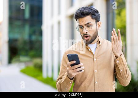 Man looking shocked while reading message on smartphone outdoors. Person with surprised expression standing outside modern building, holding phone in hand, wearing glasses and casual clothes. Stock Photo
