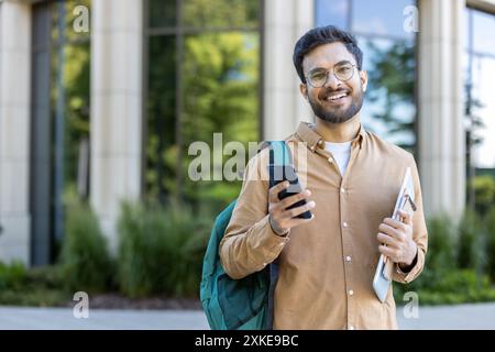 Cheerful Indian student holding phone and notebook wearing backpack standing outside modern campus building. Young man smiling, enjoying university life, casual outfit. Stock Photo