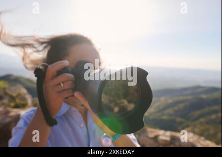 Close-up of a photographer taking pictures with a professional camera in a sunny, mountainous landscape. The wind is blowing through their hair, empha Stock Photo