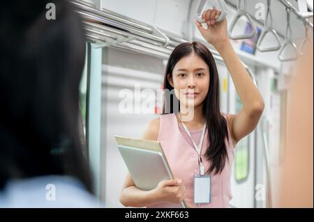 A charming, positive young Asian female office employee or businesswoman is holding a handrail and smiling at the camera while commuting by sky train Stock Photo
