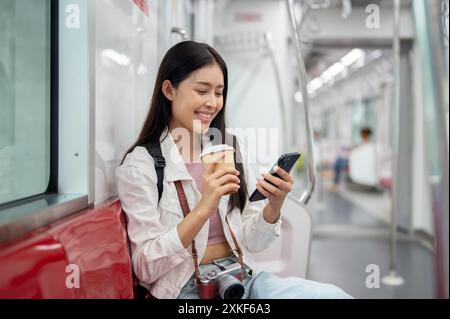 A happy, charming young Asian female tourist with a backpack is using her phone, reading text messages while taking a sky train or metro train, commut Stock Photo