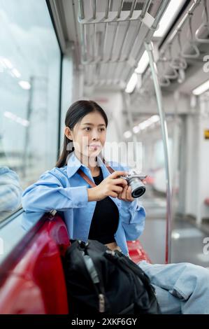 A positive young Asian female traveller is checking pictures on her camera while commuting on a sky train in the city, traveling by public transport. Stock Photo