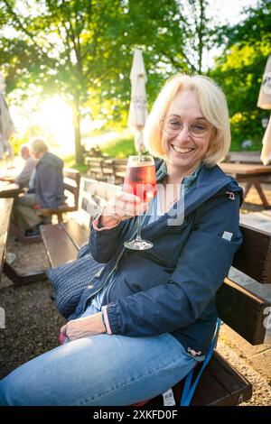 A laughing blonde woman in a beer garden in the evening backlight raises a glass of Italian aperitif Stock Photo