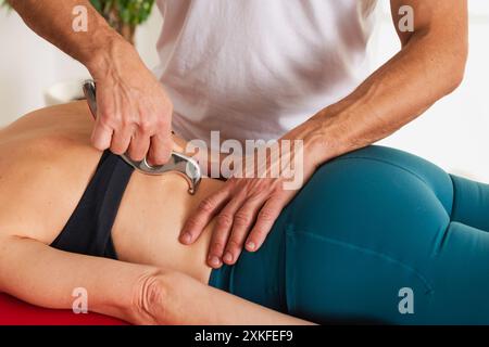 Closeup of anonymous male masseuse looking down while standing near table with faceless female client and pressing stainless IASTM tool on quadrate of Stock Photo