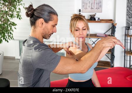 Side view of adult male with hair bun while standing observing female shoulder by holding hand for physiotherapy massage treatment in clinic with furn Stock Photo