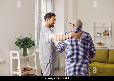 Smiling young male caregiver in uniform hugging elderly man during visit Stock Photo