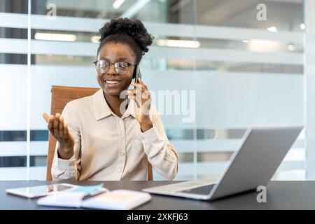 Professional businesswoman smiling while having phone conversation in modern office. Confident woman engaged in business communication, working on laptop and taking notes, Stock Photo