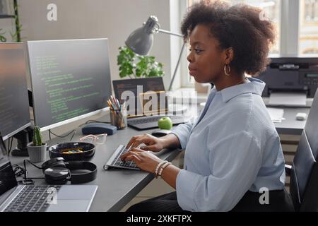 Side view of young African American female software developer using computer writing code while sitting at desk with multiple screens in office Stock Photo