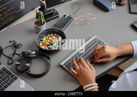 High-angle shot of gray working desk with candy bowl, headphones and hard drive placed on table while hands of female computer programmer typing source codes on keyboard in office Stock Photo