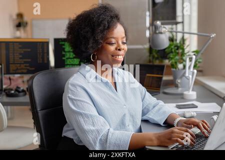 Medium portrait of young female African American computer programmer smiling typing on keyboard while sitting at desk working in IT development office, copy space Stock Photo