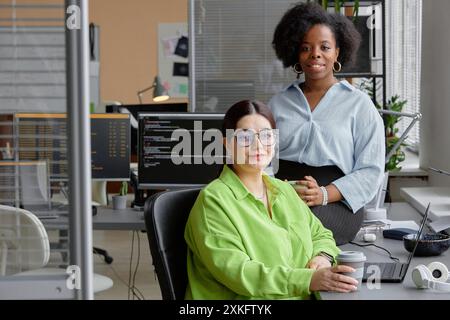 Portrait shot of two female higher-weight IT specialists on coffee break looking at camera while sitting at working desk in contemporary office with multiple computer screens, copy space Stock Photo