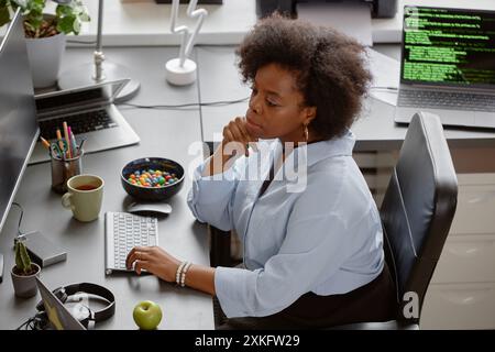 High-angle shot of thoughtful female African American senior IT specialist reading computer screen, while sitting at working desk with multiple monitors and candy bowl at hand in office Stock Photo