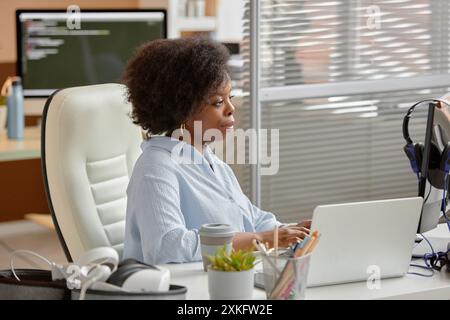 Side view portrait of young African American businesswoman sitting at desk working on computer in contemporary computer programming office. Smiling project manager typing browsing web Stock Photo