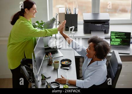 Side view of diverse group of two young female software developers cheerfully smiling giving high five to each other at working table, celebrating success in computer programmer office Stock Photo