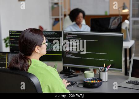 Rear view of female brunette senior IT specialist coding database script on multiple screens in software development office, copy space Stock Photo