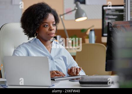 Portrait shot of concentrated young female African American software engineer typing codes looking at screen while sitting at working table in programmer office, copy space Stock Photo