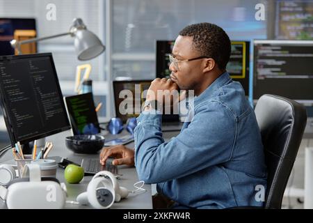 Side view of thoughtful male African American VR programmer writing code for cutting-edge game development at working desk with multiple computer screens in office, copy space Stock Photo