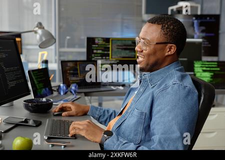 Side view shot of smiling male web developer of Black ethnicity in glasses sitting at working desk with multiple screens while using computer for code programming in office, copy space Stock Photo