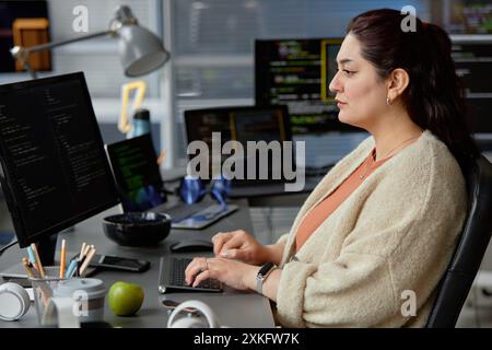 Side view shot of female Middle Eastern software engineer writing code on computer with multiple screens while sitting at desk with green apple at hand in IT company office, copy space Stock Photo