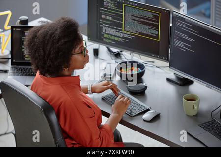 ,High-angle shot of focused curly female African American software developer at desk with candy bowl reading multiple computer screens reviewing and debugging code, while writing on keyboard in office Stock Photo