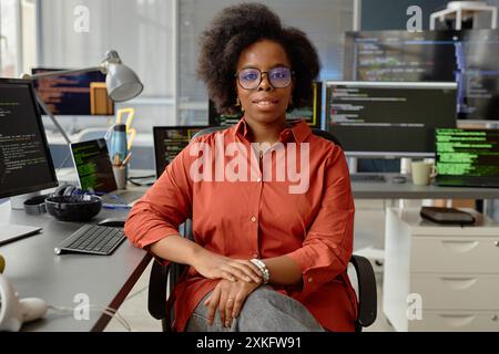 Medium portrait shot of curvy female African American VR engineer in glasses smiling looking at camera while sitting at working desk with multiple computer screens in office, copy space Stock Photo