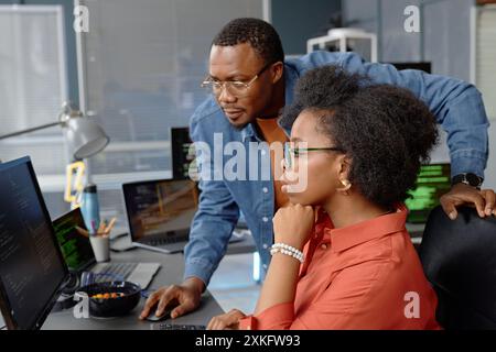 Side view shot of male senior IT specialist assisting concentrated female junior developer in reviewing code and fixing errors working on computer at desk, copy space Stock Photo