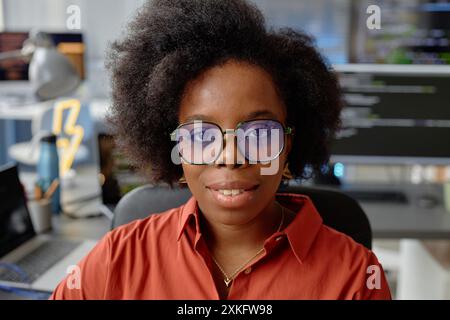 Close up portrait of curly female African American software developer wearing glasses smiling while looking at camera in computer programmer office Stock Photo