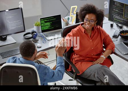 High-angle shot of smiling higher-weight female African American software engineer giving colleague high five celebrating success at large computer programmer desk in office Stock Photo
