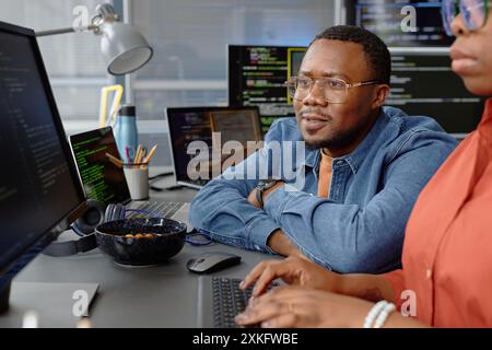 Portrait shot of pensive male African American software developer sitting at desk with folded arms reading computer screen while helping junior specialist with finding errors in code, copy space Stock Photo