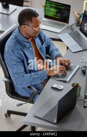 Vertical high-angle shot of concentrated male African American software developer writing program code fixing errors while sitting at working desk with laptops in office Stock Photo