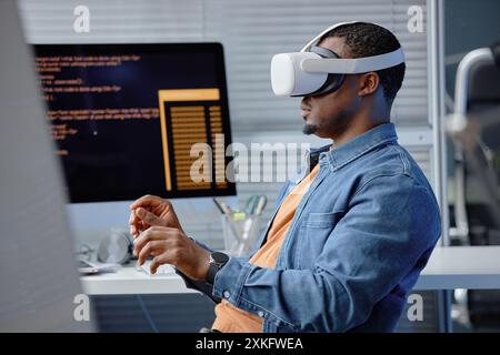 Side view shot of male African American software engineer testing VR application or video game wearing headset while sitting at computer programmer desk in office, copy space Stock Photo