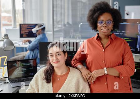 Portrait shot of two female multiethnic VR developers at working desk with computers looking at camera and smiling in IT company office Stock Photo