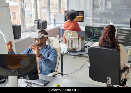 Wide angle shot of cheerful male African American software developer in headset testing VR application in office room with colleagues writing program code Stock Photo