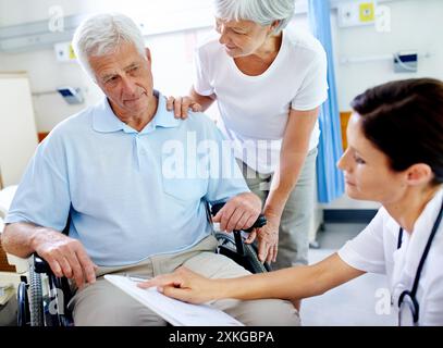Doctor, conversation and senior patient in wheelchair with a disability, happy and appointment with wife for support. Elderly man, medical worker and Stock Photo