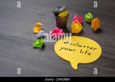 A yellow sticky note with the text "Declutter your life" on a wooden table next to a overflowing trash can and crumpled pieces of paper. Stock Photo