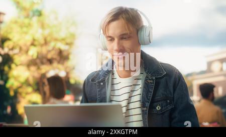 Stylish Student Working on Laptop Computer on a College Assignment Outdoors on a Terrace. Young Man Preparing for University Exams While Having a Cup of Coffee in a Cafe on a Sunny Warm Day Stock Photo