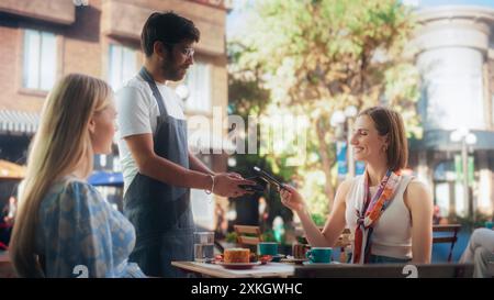Two Female Friends Ordering Delicious Cake Desserts From a Member of the Staff in a Cafe. Handsome Multiethnic Waiter Holding a Terminal, Female Using Her Smartphone for a Contactless NFC Payment Stock Photo