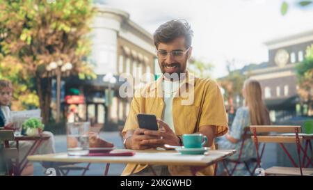 Handsome Multiethnic Man Sitting on a Terrace in a Cafe, Having a Cup of Coffee with Pastry. Happy Indian Man Connecting with Friends Online, Replying to Social Media Posts and Emails on a Smartphone Stock Photo
