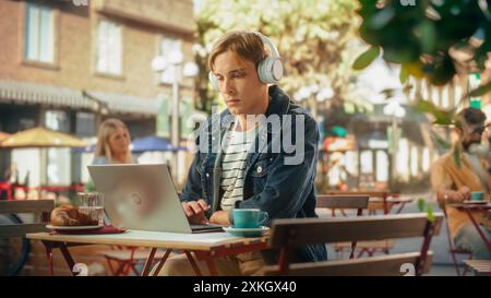 Handsome Man Using Laptop Computer on a Terrace in a Cozy Cafe on a Summer Day. Young Male Listening to Music, Drinking Espresso, Writing Work Reports and Emails Remotely Stock Photo
