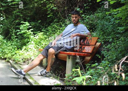 A vivid image featuring a young tattooed man in sporting clothes, looking bored and waiting on a bench on a sunny day in the city of Zurich. Stock Photo