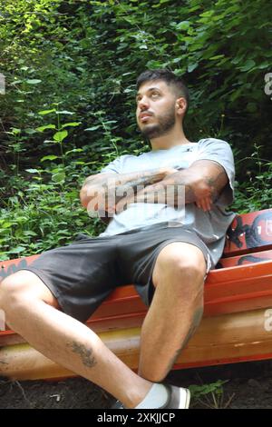 A vivid image featuring a young tattooed man in sporting clothes, looking bored and waiting on a bench on a sunny day in the city of Zurich. Stock Photo