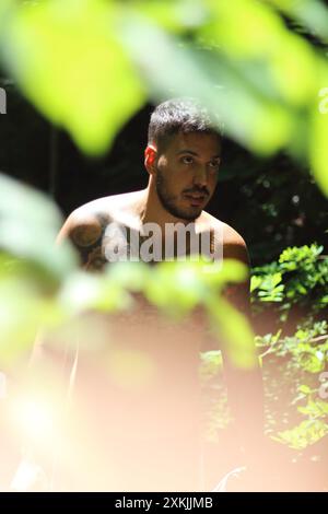 A young, tattooed, and muscular man stands framed by lush green leaves on a sunny day in Zurich. The cityscape backdrop adds a vibrant urban touch to Stock Photo