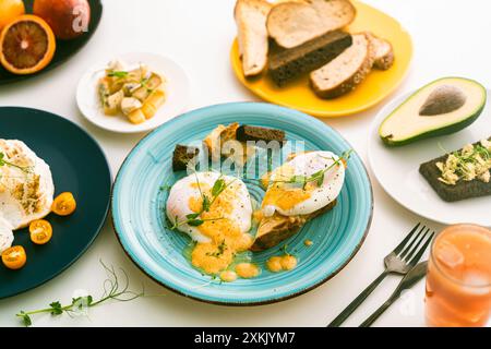 Eggs Benedict with hollandaise sauce and peas on a turquoise plate and avocado with rye toast, cheese, blood oranges and orange juice on a white table Stock Photo