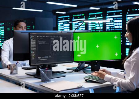 Colleagues working on green screen desktop PC in data center housing storage infrastructure. Coworkers using mockup computer display in server hub workspace filled with networking tech Stock Photo