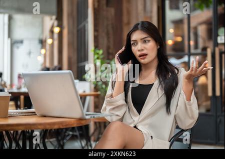 A moody, dissatisfied Asian businesswoman is talking on the phone with her assistant, complaining about work while working remotely at a cafe in the c Stock Photo