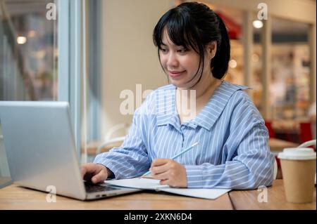 A young beautiful Asian woman is studying online via her laptop or working remotely from a cafe, looking at the laptop screen. city life, lifestyle, t Stock Photo