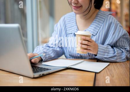 A cropped image of a happy young Asian woman is having coffee while studying an online class, working remotely from a coffee shop. people and technolo Stock Photo