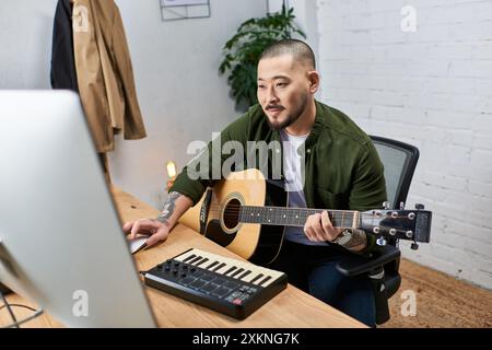A young Asian man plays an acoustic guitar and uses a keyboard in his studio. Stock Photo